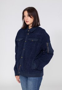 Granatowa kurtka z teksturowanym, puszystym wykończeniem i gładkimi akcentami z materiału. Posiada przednie kieszenie, zapięcia na guziki oraz kieszeń na suwak na rękawie.