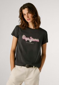 T-shirt de algodão preto com estampa do logotipo em rosa e branco "Pepe Jeans LONDON". Decote redondo, mangas curtas, combinado com calças bege e um cinto.