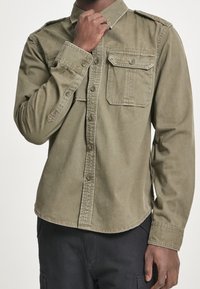 Olivgrön långärmad skjorta i bomull, med knäppning framtill, två bröstfickor och axelklaffar. Slät textur, rundad nederkant.