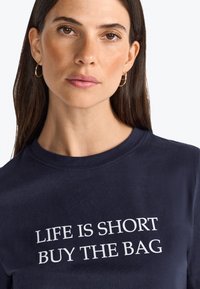 T-shirt en coton bleu marine à manches courtes, avec un texte blanc qui dit "LA VIE EST COURTE ACHÈTE LE SAC" sur la poitrine.
