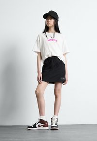 Vit t-shirt med rosa "JORDAN" tryck, svarta shorts med logotyp, svart keps och vita strumpor med svarta och röda högstående sneakers.