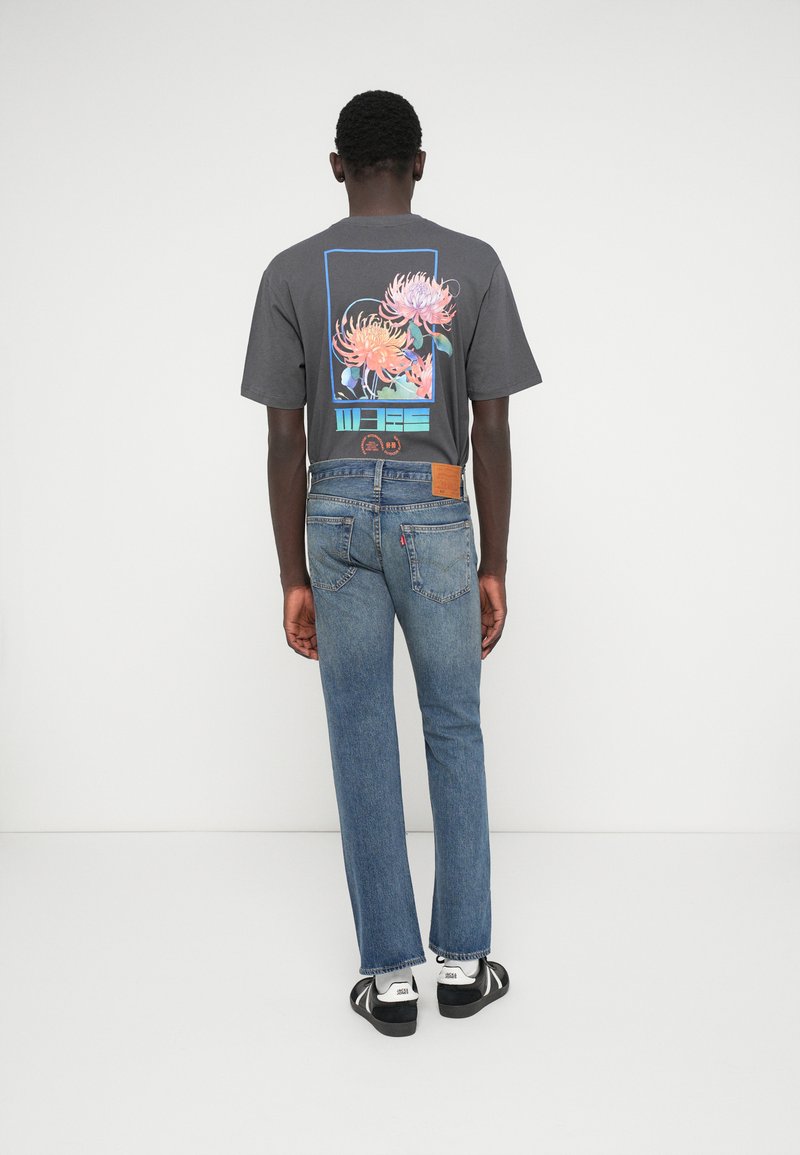 Camiseta gráfica gris con diseño de flores en la parte posterior; combinada con jeans rectos azules. Se llevan zapatillas blancas y negras.