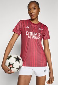 Κόκκινη αθλητική φανέλα με ζιγκ-ζαγκ σχέδιο, με το λογότυπο «Emirates» σε λευκό. Συνδυάζεται με λευκό σορτς και μια ασπρόμαυρη ποδοσφαιρική μπάλα.