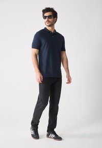 Polo navy de manga corta, con un logo sutil, combinado con jeans negros y zapatillas negras con rayas blancas. Los accesorios incluyen gafas de sol.