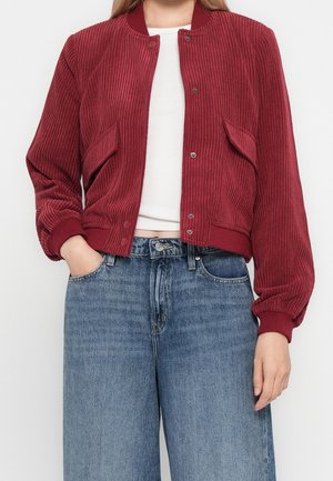 Person iført en rød ribstrikket jakke over en hvid top og blå højttaljede vidde jeans med den ene hånd i lommen.