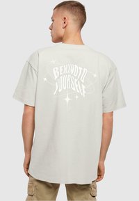 Lichtgrijze oversized t-shirt van katoen, met een opvallende witte grafische afbeelding op de achterkant met de tekst "WEES VRIENDELIJK VOOR JOUWZELF" en sterren.
