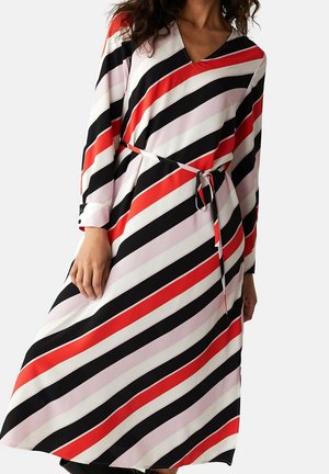 Kobieta ubrana w długą sukienkę z długim rękawem, z paskiem, w ukośne paski w kolorach czerwonym, czarnym, białym i jasnoróżowym na jednolitym tle.