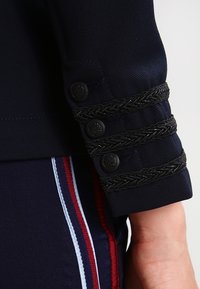 Manica di un blazer blu navy con tre dettagli intrecciati neri e quattro bottoni tondi. Texture liscia con una vestibilità su misura.
