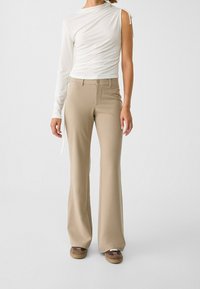 Vrouw gekleed in beige high-waist flare broeken, een witte one-sleeve ruches top en bruine schoenen, staand tegen een effen lichte achtergrond.