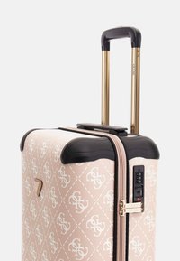 Valise rigide rose avec motif de logo blanc, accents noirs contrastants et poignée en métal doré. Elle dispose d'un verrou à combinaison et d'une fermeture éclair.