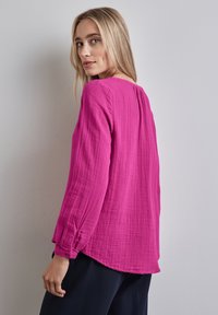 Blusa fucsia a maniche lunghe con tessuto testurizzato, vestibilità comoda e polsini con bottoni. Presenta un collo rotondo e dettagli plissettati sul retro.