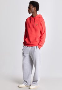 Sweatshirt rouge à capuche avec une poche avant et un logo jaune. Associé à un pantalon de jogging gris, ample et à des chaussures de couleur claire.