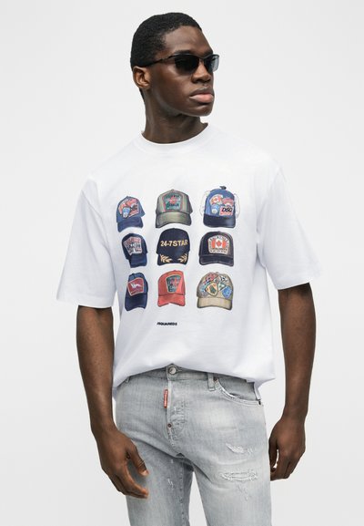 Άνδρας που φοράει μαύρα γυαλιά ηλίου, λευκό T-shirt με εννέα πολύχρωμες εικονογραφήσεις από καπέλα του μπέιζμπολ, ανοιχτό γκρι τζιν με φθορές, μπροστά σε απλό φόντο.