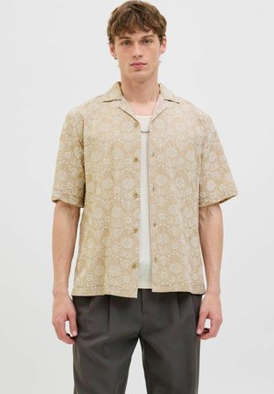 Chemise beige à manches courtes avec des motifs floraux, en tissu léger, avec une fermeture à boutons et une coupe décontractée, associée à un sous-vêtement blanc texturé et un pantalon gris.