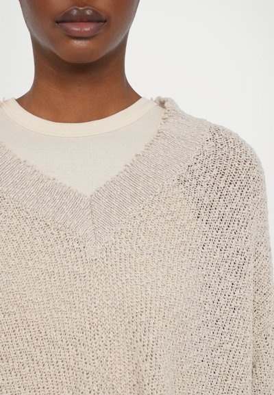 Knit sweater in light beige with a v-neckline. Features a loose weave texture and frayed edges along the collar. Layered over a ribbed shirt.