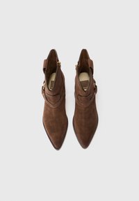 Botas de tobillo de ante marrón con puntas afiladas, detalles decorativos de correa y cremalleras laterales. Textura suave con un acabado mate.
