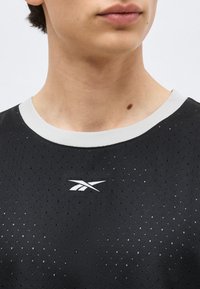 Κοντινή λήψη του λαιμού και του στήθους ενός ατόμου που φοράει μαύρο διατρητό αθλητικό μπλουζάκι Reebok με λευκό γιακά και λογότυπο στο κέντρο κάτω από τον λαιμό.