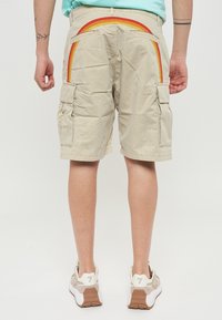 Pantaloni cargo beige con un motivo arcobaleno sulle tasche posteriori, dotati di due tasche laterali e una vestibilità comoda. Indossati con sneakers dai toni neutri.
