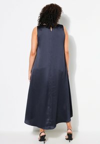 Gaun maxi tanpa lengan berwarna navy dengan tekstur halus, lipatan halus, dan detail lubang kunci kecil di bagian belakang. Dipadukan dengan sandal hak tinggi hitam.