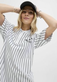 Jeune femme avec des cheveux blonds portant une casquette noire et un polo blanc avec des rayures verticales noires, posant les mains derrière la tête.