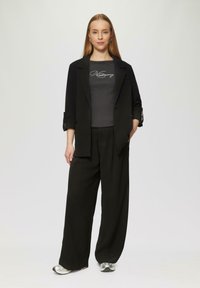 Schwarzes Blazer mit hochgekrempelten Ärmeln über einem taillierten grauen Oberteil mit silberner Schrift. Weit geschnittene schwarze Hose und silberne Sneakers runden das Outfit ab.