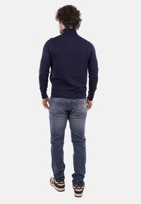 Maglione blu navy con collo e polsini a coste, abbinato a jeans in denim chiaro e sneakers marroni, visto da dietro.