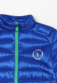 Modrá péřová bunda s předním zipem a zeleným zipem, s kovovým logem a designem v švu ve tvaru diamantů.