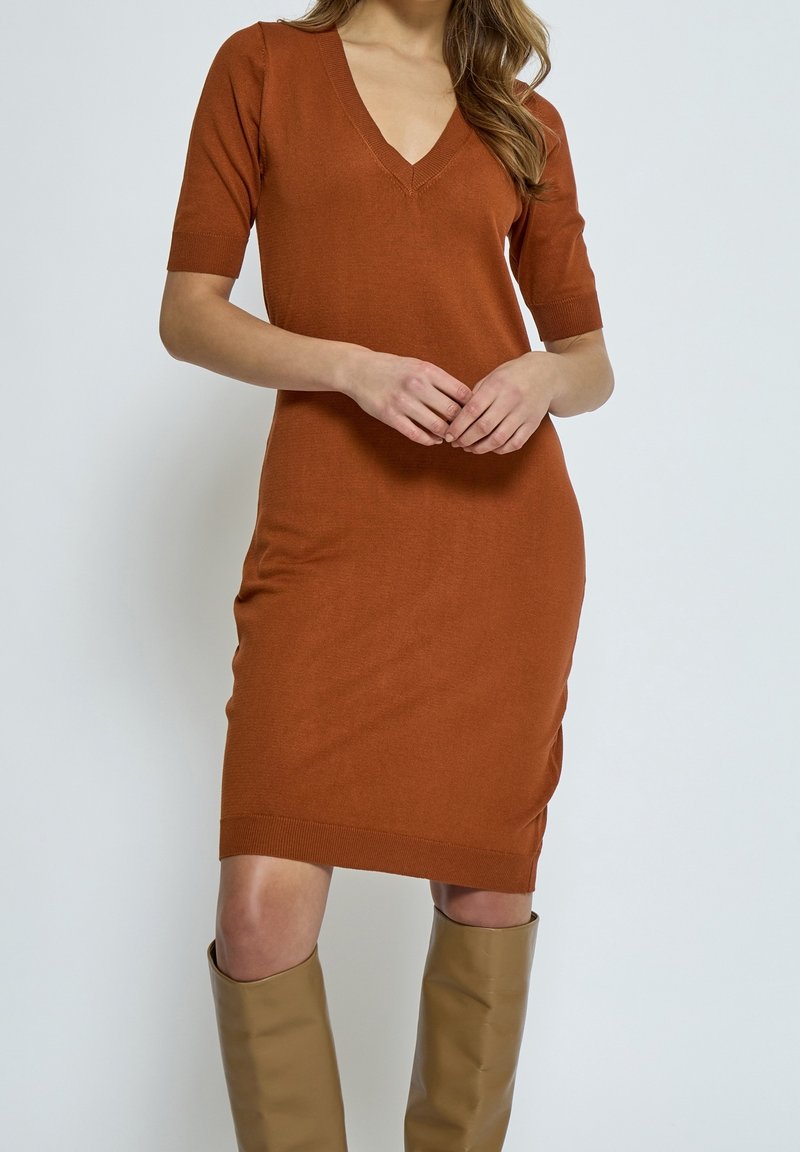 Robe tricotée couleur rouille avec un décolleté en V et des manches courtes. Coupe ajustée, longueur mi-cuisse et accents de bord côtelé. Texture lisse.