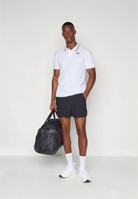 Fehér galléros póló piros díszítéssel a galléron, fekete rövidnadrág, fehér tornacipő és egy fekete utazótáska. Egyszerű, hétköznapi sportos viselet.