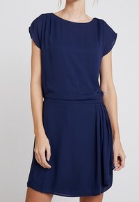 Robe courte bleu marine en tissu léger, présentant des manches courtes, une taille cintrée et un détail plissé sur un côté.