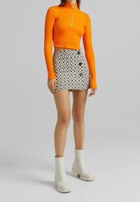 Orange ribbad långärmad crop top med hög krage, ihop med en mönstrad svart och vit minikjol med knapppå detaljer och vita robusta ankelskor.