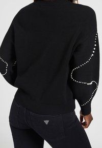 Schwarzer Strickpullover mit Perlenverzierungen an den Ärmeln, mit einem lockeren Schnitt und geripptem Saum. Kombiniert mit schwarzen Jeans, die ein Logo-Patch zeigen.