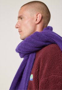 Vijoličasti šal iz mehke, teksturirane tkanine, ovit okoli vratu, v kombinaciji z burgonjsko flis jakno z barvitim logotipom.