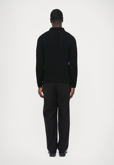 Pull noir en tricot avec un motif en câble texturé et un ourlet côtelé. Porté avec un pantalon sombre. Col classique au niveau de l'encolure.