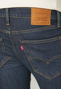 Jeans de denim azul escuro com um patch de marca em pele, costura amarela e uma etiqueta vermelha no bolso traseiro. Corte slim com um design de perna afunilada.
