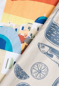 Δύο διπλωμένα υφάσματα, το ένα με πολύχρωμα κτίρια ελληνικών νησιών και μπλε τρούλους, το άλλο με μπλε ψάρια και εικόνες φέτας λεμονιού σε μπεζ φόντο.