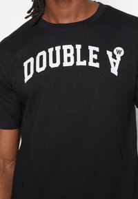 Černé bavlněné tričko s bílým, tučným textem "DOUBLE V" v obloukovém formátu. Obsahuje malou kulatou nášivku s detailem "W".