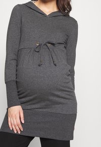 Šedý tunikový svetr s kapucí, dlouhými rukávy, stahováním v pase a žebrovaným lemem. Vyrobený z měkké látky, navržený pro pohodlný střih přes těhotenské bříško.