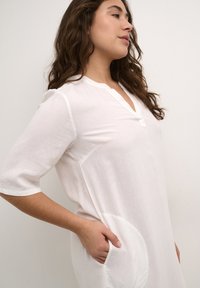 Robe blanche légère avec une texture douce, dotée d'un col en V, de manches courtes et de poches latérales aux bords arrondis.