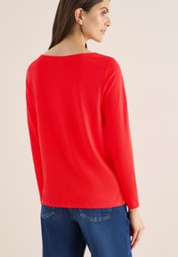 Maglietta rossa a maniche lunghe con scollatura ampia, texture liscia e vestibilità morbida, abbinata a jeans in denim blu. Design semplice, senza motivi o dettagli.