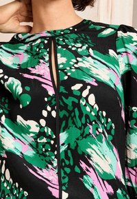 Blusa multicolore con motivi astratti verdi, rosa e bianchi su sfondo nero, caratterizzata da un colletto a chiave e tessuto testurizzato.