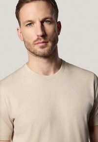 Beżowa koszulka z krótkim rękawem i okrągłym dekoltem, wykonana z miękkiego materiału i bezszwowych ramion, o gładkiej fakturze.