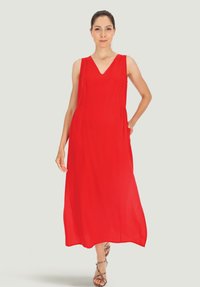 Czerwona długa sukienka maxi z dekoltem w kształcie litery V, bez rękawów i z bocznymi kieszeniami. Lekka tkanina o gładkiej strukturze. Modelka nosi sandały na paski.