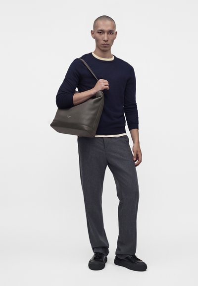 Homme portant un pull bleu marine, un pantalon gris et des chaussures noires, tenant un grand sac à bandoulière en cuir gris.