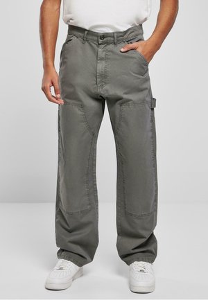 Mann trägt lockere graue Cargo-Hosen mit seitlichen und Knie-Taschen, weiße Turnschuhe und ein weißes T-Shirt, steht mit den Händen in der Nähe der Taschen.