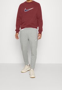 Sudadera Nike burdeos con logo blanco y azul marino, combinada con pantalones de chándal gris claro que presentan un pequeño logo blanco, complementada con zapatillas beige.