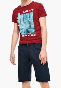 Muž v červeném grafickém tričku s modrým vlnkovým potiskem a tmavě modrých šortkách po kolena, stojící v uvolněné pozici.