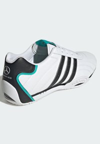 Biele atletické tenisky s koženým zvrškom, čiernymi a tyrkysovými akcentmi, tromi čiernymi pruhmi a textúrovanou gumovou podrážkou. Obsahujú logo Mercedes-Benz.