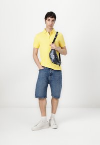 Gelbes Poloshirt, blaue Denim-Shorts, weiße Sneaker, graue Schultertasche mit Muster. Modell steht vor einem schlichten weißen Hintergrund.