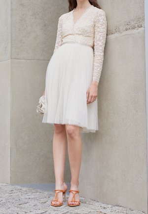 Hellbeiges Kleid mit langen Spitzenärmeln, einem V-Ausschnitt und einem Tüllrock. Kombiniert mit orangenen Riemchenabsätzen. Hält eine kleine, helle Clutch.